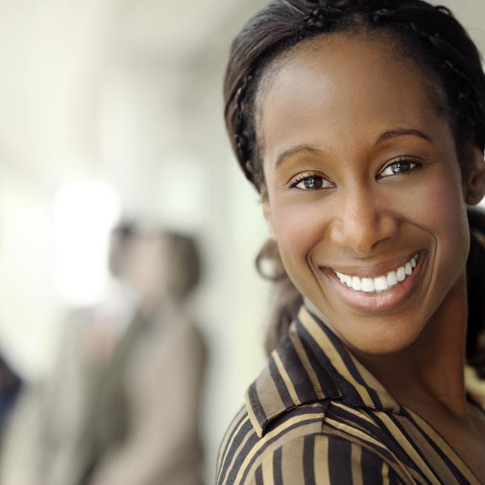 A woman smiles whilst a blurry small group of people in business wear stand in conversation in the background